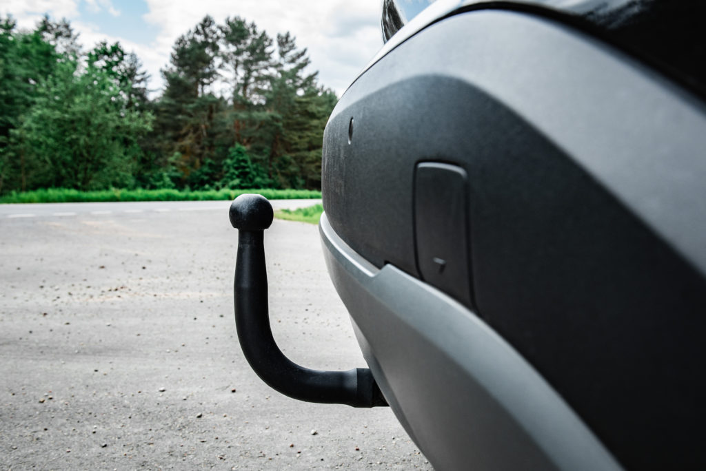 Tył samochodu z widocznym czarnym hakiem holowniczym zamontowanym pod zderzakiem, zaparkowanym przy drodze w otoczeniu zieleni.