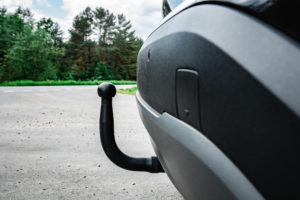 Tył samochodu z widocznym czarnym hakiem holowniczym zamontowanym pod zderzakiem, zaparkowanym przy drodze w otoczeniu zieleni.